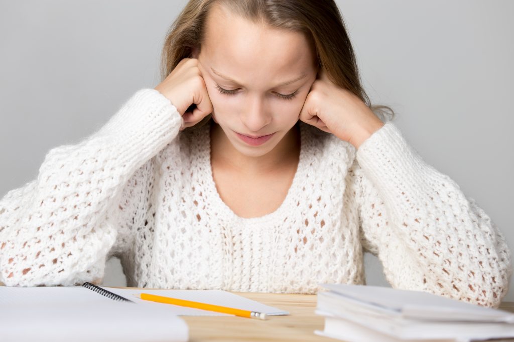 Portrait of stressed beautiful casual girl, sitting at desk, holding her head in arms with exhausted frustrated expression, cannot understand hard school task, studio, gray background problèmes practical